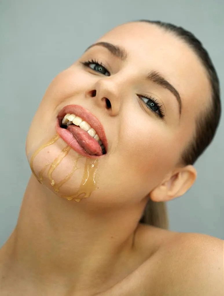 Close-up of a smiling woman with honey dripping down her chin, licking her lips playfully. 