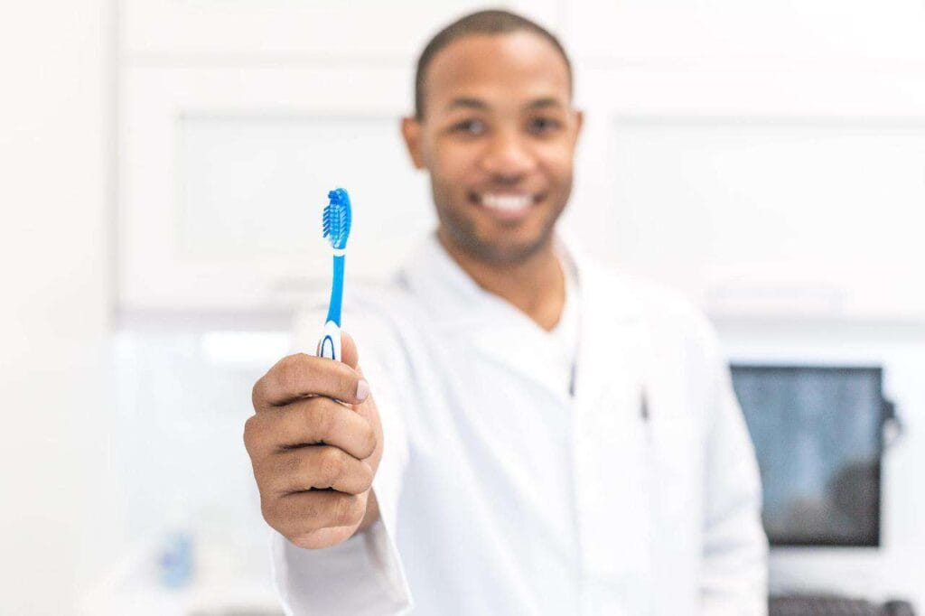 Dentist holding a blue toothbrush towards the camera in a clinical setting.