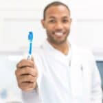 Dentist holding a blue toothbrush towards the camera in a clinical setting.