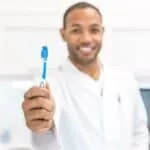 Dentist holding a blue toothbrush towards the camera in a clinical setting.