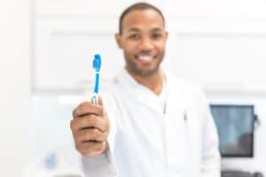 Dentist holding a blue toothbrush towards the camera in a clinical setting.