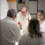 Older couple in white bathrobes brushing their teeth together in front of a bathroom mirror.