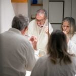 Older couple in white bathrobes brushing their teeth together in front of a bathroom mirror.