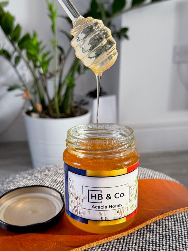 A jar of HoneyBee & Co. Acacia Honey with a honey dipper, showcasing its golden colour and smooth texture. Pure raw British and European honey available from your local honey shop in the UK.