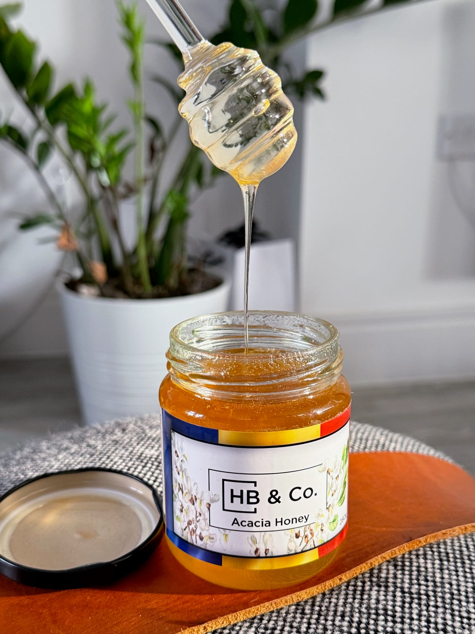 A jar of HoneyBee & Co. Acacia Honey with a honey dipper, showcasing its golden colour and smooth texture. Pure raw British and European honey available from your local honey shop in the UK.