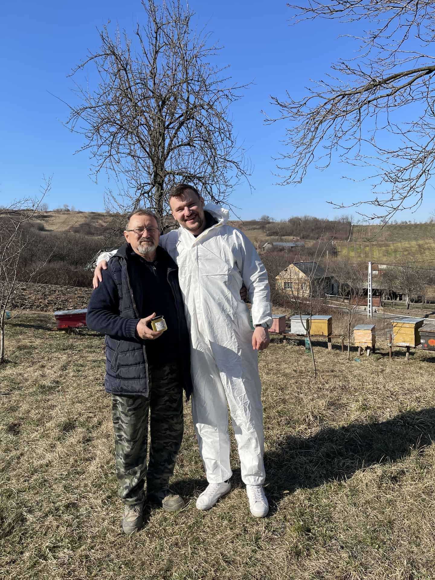 Our Founders. Nistor Fanel and Nistor Dragos. Two Romanian beekeepers standing together near colourful hives in the countryside, representing HoneyBee & Co.’s partnership with local beekeepers and sustainable honey sourcing.