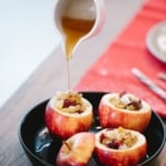 Warm honey being drizzled from a jug over baked stuffed apples filled with nuts and dried fruit, served on a black plate.