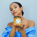 Woman holding a jar of raw honey in front of a light background, wearing a blue off-shoulder top.