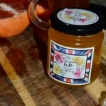 A jar of HoneyBee & Co. Wild Flower honey with a floral Union Jack label, sitting on a rustic wooden surface next to a sliced orange.