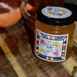 A jar of HoneyBee & Co. Wild Flower honey with a floral Union Jack label, sitting on a rustic wooden surface next to a sliced orange.
