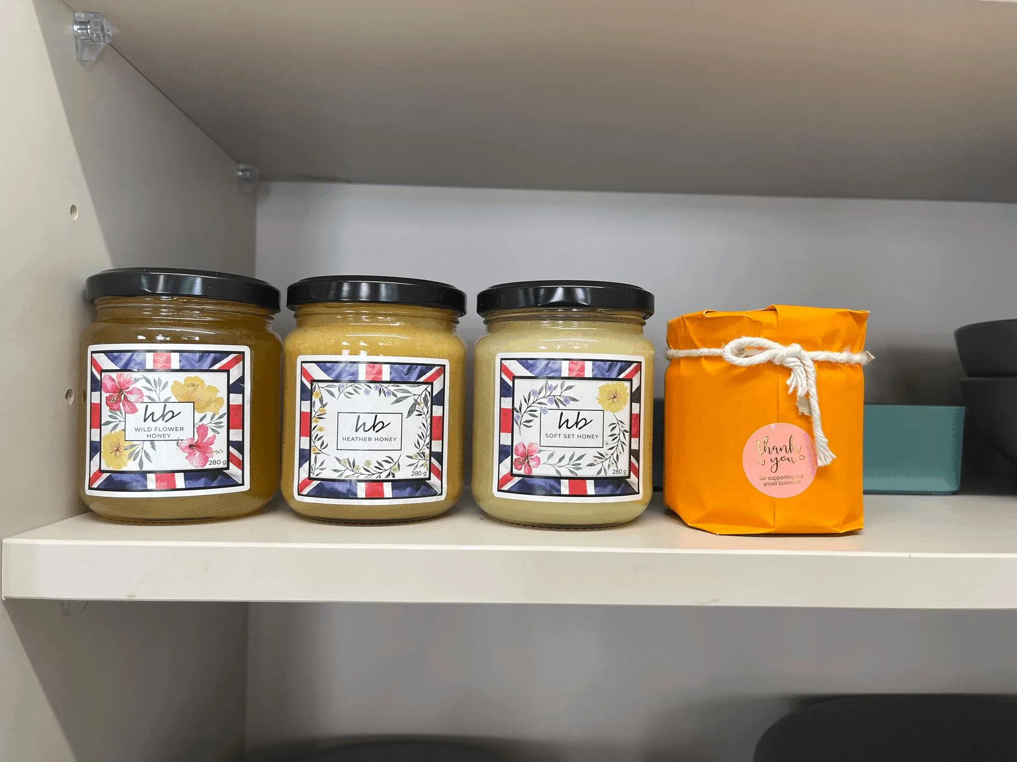 Three jars of HoneyBee & Co. Union Jack edition honey and one bright orange-wrapped gift jar displayed on a white shelf.