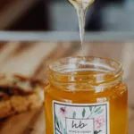Solid State of Raw Honey Acacia Honey A spoon drizzling raw golden honey into a glass jar with a botanical HoneyBee & Co. label, placed on a wooden table.