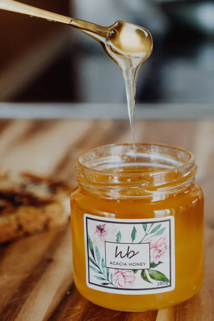 Solid State of Raw Honey Acacia Honey A spoon drizzling raw golden honey into a glass jar with a botanical HoneyBee & Co. label, placed on a wooden table.