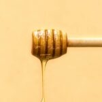 Close-up of a honey dipper with golden honey dripping against a warm yellow background.