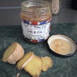A jar of HoneyBee & Co. Heather Honey with slices of fresh ginger on a dark green surface, highlighting its thick texture and rich golden colour.