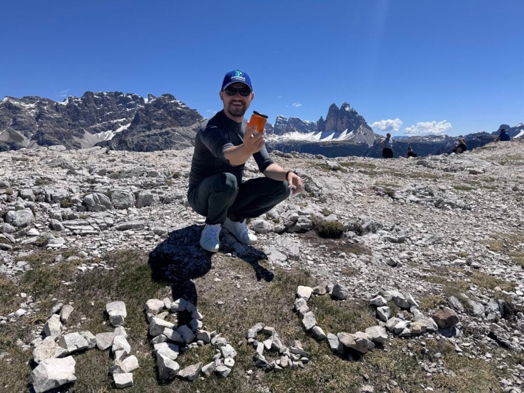 Syrups & Sweeteners: Raw Honey 1 Person sitting on a rocky mountain plateau under a clear blue sky, holding a small object with dramatic alpine peaks in the background